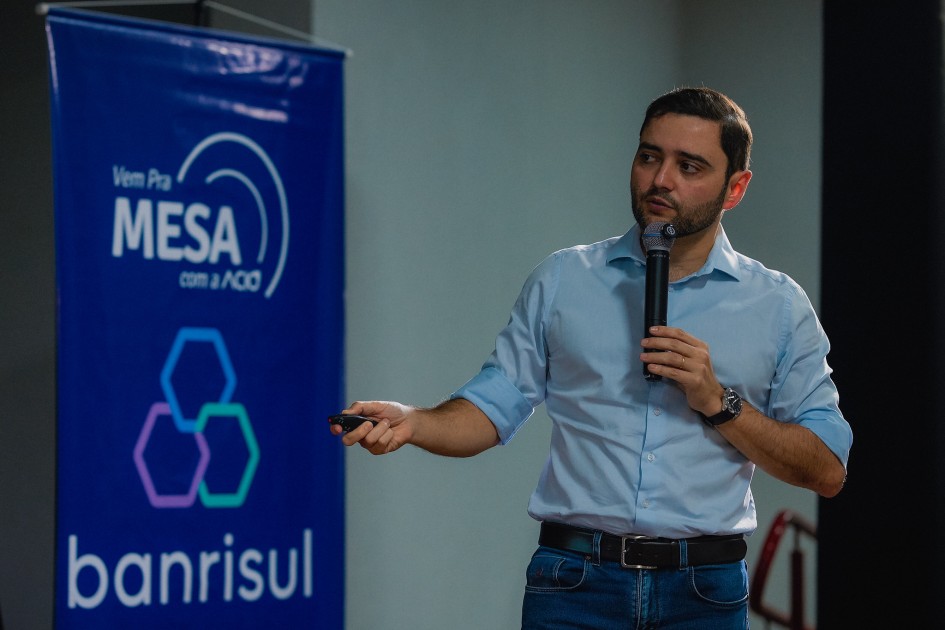 Gabriel aparece em close, segurando um microfone e apontando com a outra mão. Atrás dele, o banner azul do Banrisul com a frase “Vem Pra Mesa com a ACI”.