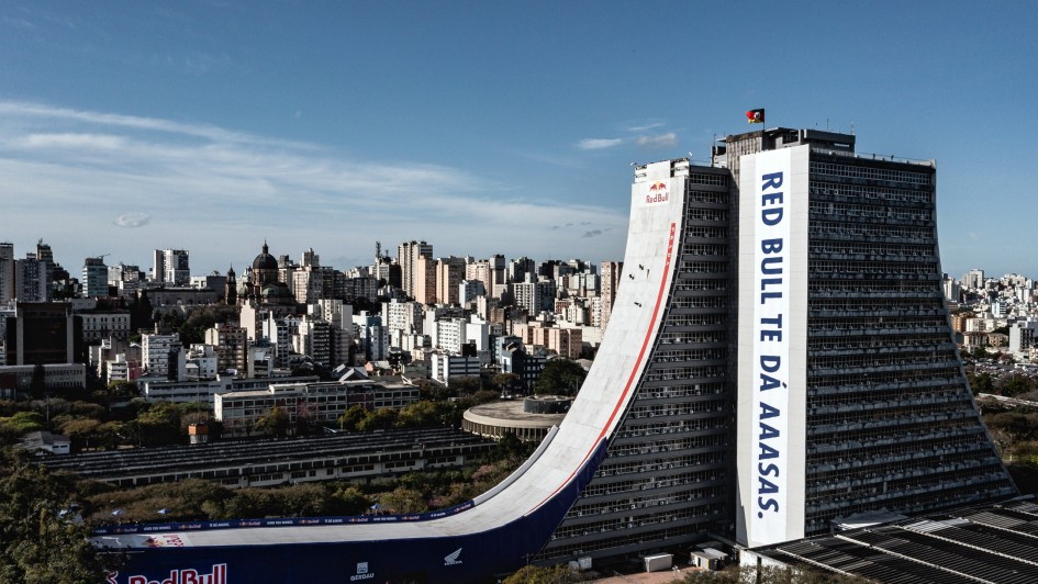 A imagem mostra um grande edifício em área urbana com uma estrutura arquitetônica incomum: uma enorme rampa que se estende do topo até o chão. A rampa é personalizada com logotipos e frases da marca Red Bull, incluindo "RED BULL TE DÁ AAASAS". Ao fundo, vê-se uma paisagem urbana densa, com diversos prédios altos.
