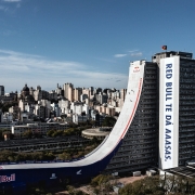 A imagem mostra um grande edifício em área urbana com uma estrutura arquitetônica incomum: uma enorme rampa que se estende do topo até o chão. A rampa é personalizada com logotipos e frases da marca Red Bull, incluindo "RED BULL TE DÁ AAASAS". Ao fundo, vê-se uma paisagem urbana densa, com diversos prédios altos.