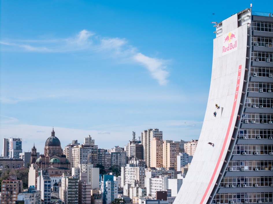 A imagem mostra uma paisagem urbana com destaque para um edifício que possui uma grande rampa curva em sua lateral, marcada com o logotipo da Red Bull no topo. A rampa está sendo usada para atividades de skate. Ao fundo, há diversos prédios e estruturas arquitetônicas.