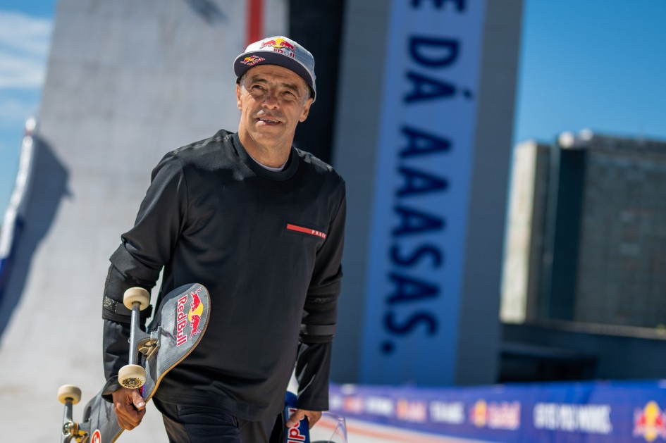 A imagem mostra um homem segurando um skate, vestindo uma camiseta preta de manga longa e calça escura, com um boné da Red Bull.
