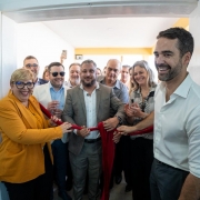 Um grupo de pessoas participa de uma cerimônia de inauguração em ambiente interno, de frente para uma porta branca aberta. Ao centro, uma mulher de blazer amarelo-mostarda e óculos de armação grossa azul segura uma tesoura grande e sorri, pronta para cortar a faixa vermelha que simboliza a abertura oficial. Do lado direito, um homem de camisa branca de mangas dobradas também segura a faixa e sorri amplamente. Atrás deles, várias pessoas, homens e mulheres, acompanham a cena, algumas batendo palmas e outras registrando o momento com celular. O clima é festivo e descontraído.