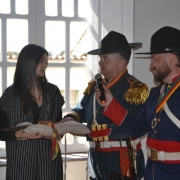 Dois homens vestidos com trajes históricos de inspiração militar farroupilha, incluindo fardas azuis com detalhes vermelhos, ombreiras douradas e chapéus de aba larga, participam de uma solenidade em ambiente interno. Um deles segura um microfone enquanto o outro observa. À esquerda, uma mulher de cabelos longos e escuros veste roupa preta com sobreposição listrada e segura uma almofada com uma lança histórica apoiada, envolta em uma faixa colorida nas cores do Rio Grande do Sul. Ao fundo, há uma janela ampla iluminando a cena.
