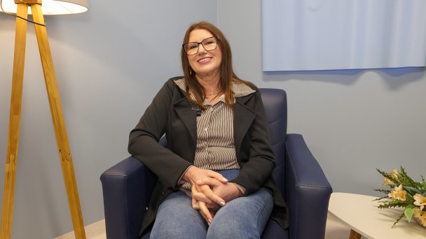 Uma mulher de óculos, cabelos castanhos e pele clara sorri para a câmera. Ela está sentada em uma poltrona de cor azul escuro. Ao lado esquerdo, há uma luminária e, ao lado direito, uma mesinha com flores. 