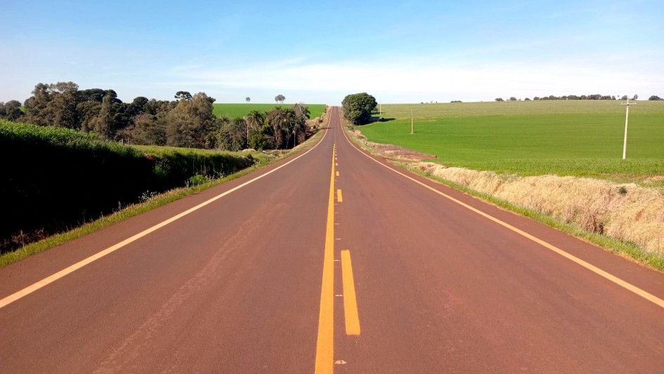 Descrição para fins de acessibilidade: Fotografia de uma estrada asfaltada em linha reta, com duas faixas e marcações amarelas no centro e nas laterais. A via atravessa uma paisagem rural, com plantações verdes à direita e árvores mais altas à esquerda. Ao fundo, a estrada segue em direção ao horizonte, sob um céu azul claro sem nuvens.