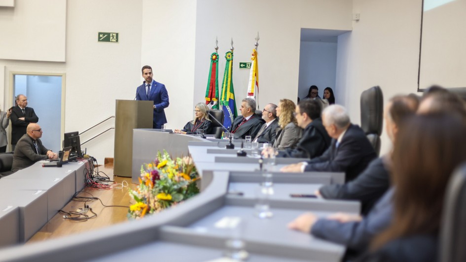 Leite está de terno azul e em pé, falando em um púlpito em um auditório. Na mesa em frente a ele, várias pessoas estão sentadas em cadeiras altas, ouvindo a fala. Há bandeiras do Brasil e do Rio Grande do Sul ao fundo, além de arranjos de flores na frente da mesa.