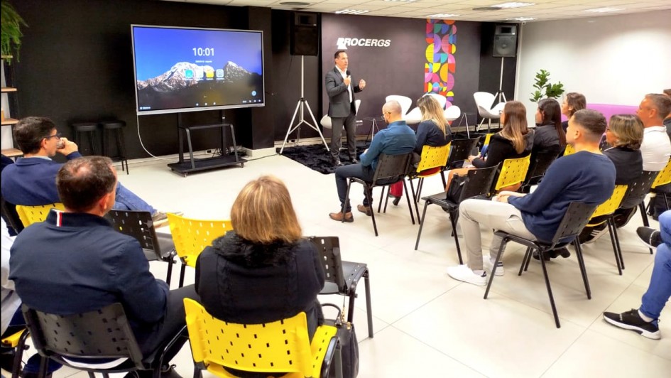 Na imagem, um homem em traje formal fala em pé diante de um grupo de pessoas sentadas em cadeiras pretas e amarelas organizadas em fileiras. Ele está posicionado próximo a um telão. O ambiente é uma sala moderna, bem iluminada, com teto de placas brancas, paredes escuras e o logotipo “PROCERGS” em destaque.