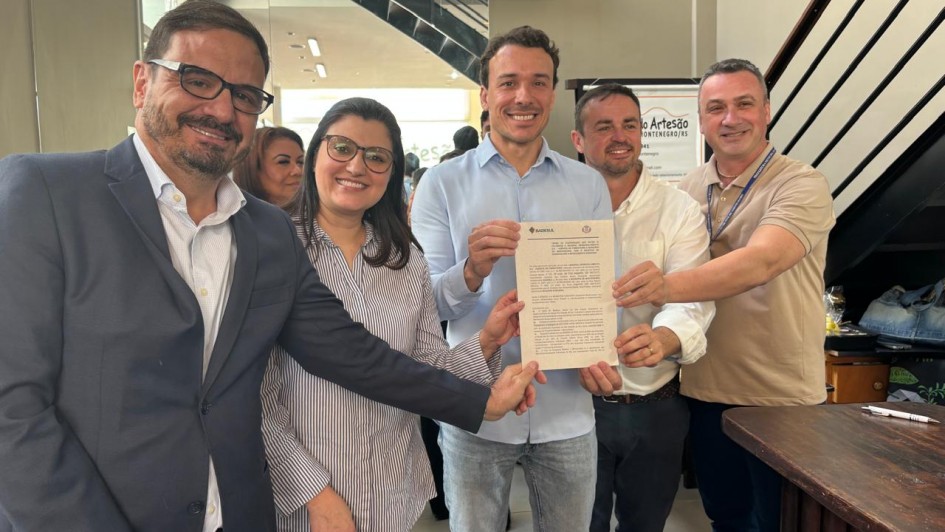 Cinco pessoas estão em pé, sorrindo, posando para a foto enquanto seguram juntas um documento oficial. Estão em um ambiente interno, bem iluminado, com decoração moderna. À esquerda, um homem de óculos, barba e terno azul escuro. Ao lado dele, uma mulher de óculos, com cabelo castanho e camisa listrada. No centro, um homem de camisa azul clara segura o documento com as duas mãos. À sua direita, outro homem de camisa branca e, ao lado, um homem de camisa polo bege com crachá. Ao fundo, há um espelho e um cartaz com os dizeres parcialmente visíveis “...Artesão... Montenegro/RS”. Todos estão participando de um evento oficial, relacionado ao lançamento de um programa governamental.