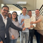 Cinco pessoas estão em pé, sorrindo, posando para a foto enquanto seguram juntas um documento oficial. Estão em um ambiente interno, bem iluminado, com decoração moderna. À esquerda, um homem de óculos, barba e terno azul escuro. Ao lado dele, uma mulher de óculos, com cabelo castanho e camisa listrada. No centro, um homem de camisa azul clara segura o documento com as duas mãos. À sua direita, outro homem de camisa branca e, ao lado, um homem de camisa polo bege com crachá. Ao fundo, há um espelho e um cartaz com os dizeres parcialmente visíveis “...Artesão... Montenegro/RS”. Todos estão participando de um evento oficial, relacionado ao lançamento de um programa governamental.