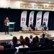 Em um auditório lotado, uma mulher está de pé no palco, falando ao microfone. Há quatro poltronas ao lado, com duas ocupadas por um homem e uma mulher sentados. Ao fundo, há banners que remetem à doação de órgãos, com a marca do governo do Estado. 