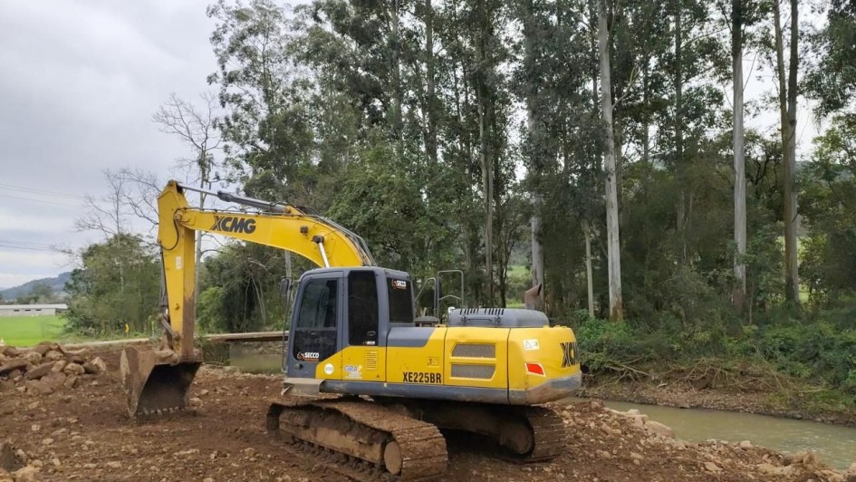 Fotografia de uma escavadeira hidráulica amarela, modelo XE225BR da marca XCMG, estacionada sobre um terreno de terra e pedras às margens de um rio. Ao fundo, há uma área de mata com árvores altas e de troncos claros, além de vegetação densa. O céu está nublado, compondo um cenário de trabalho em área natural.