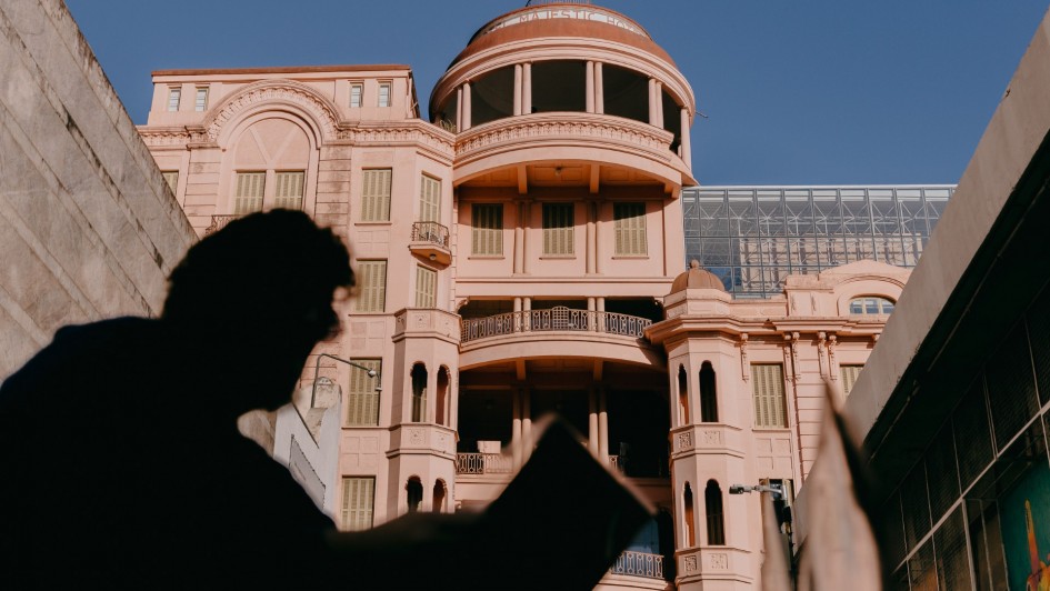 Na imagem, a fachada rosa do prédio histórico da Casa de Cultura Mario Quintana (antigo Hotel Majestic) ocupa o fundo sob céu azul. Em primeiro plano, aparece a silhueta desfocada de uma pessoa segurando um livro aberto, criando contraste entre sombra e luz.