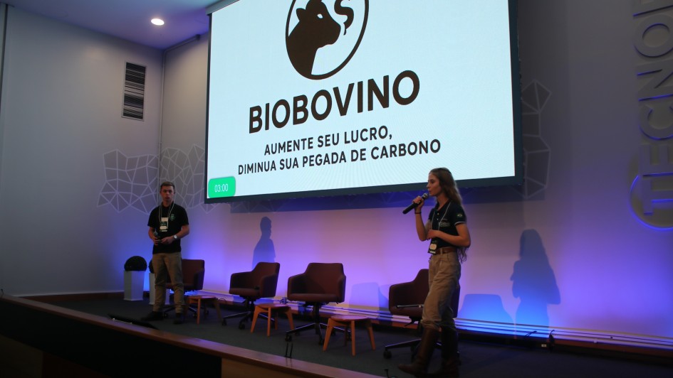 Descrição da imagem para fins de acessibilidade:
Duas pessoas apresentam no palco de um auditório. À esquerda, um homem de camiseta preta e calça bege segura um controle. À direita, uma mulher com camiseta preta, calça bege e botas marrons fala ao microfone. No telão atrás deles, aparece o logotipo de um boi com um cifrão, seguido do nome BIOBOVINO e do slogan: “Aumente seu lucro, diminua sua pegada de carbono”. Há três cadeiras e duas mesinhas no palco. O ambiente é moderno, com iluminação azulada.