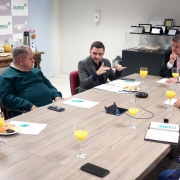 Reunião em uma sala corporativa, com cerca de sete homens sentados ao redor de uma mesa retangular de madeira clara. No centro da mesa estão taças com suco de laranja, pratos pequenos com salgados e frutas, além de cadernos, papéis e canetas. Um dos participantes, no centro da imagem, fala gesticulando com as mãos, enquanto os demais ouvem com atenção. Ao fundo, há um aparador com mais alimentos e bebidas, e um painel com a logomarca “AMRIGS” repetida em padrão. Na parede lateral, são visíveis certificados e placas de premiação em exposição. A cena transmite um ambiente formal de reunião e diálogo institucional.