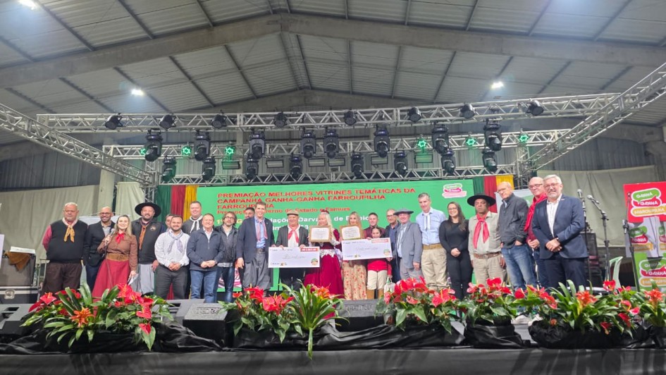 Na imagem, um grupo de pessoas posa no palco durante cerimônia de premiação da campanha Ganha-Ganha Farroupilha. Homens e mulheres, alguns em trajes típicos gaúchos, estão alinhados em frente ao painel com a identidade visual da campanha. Entre eles, participantes exibem certificado e cheque simbólico, enquanto o cenário inclui iluminação de palco e arranjos florais vermelhos à frente.