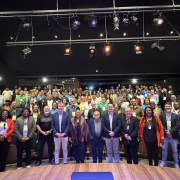 A imagem mostra uma foto de grupo tirada em um auditório com iluminação de palco. Há aproximadamente 100 pessoas presentes, distribuídas entre o palco (na frente) e as poltronas do auditório (ao fundo). Na primeira fileira, à frente, estão cerca de 20 pessoas em pé, possivelmente autoridades, organizadores ou convidados de destaque. Entre elas, há homens e mulheres de diferentes idades e etnias, vestidos de forma formal e semiforma, alguns com crachás de identificação pendurados no pescoço.
Ao fundo, o restante do grupo ocupa as cadeiras do auditório, posando para a foto coletiva. As luzes de palco estão acesas, iluminando a cena, e é possível ver refletores e equipamentos de iluminação no teto. O clima geral é de celebração ou encerramento de um evento importante, possivelmente relacionado à cultura ou à gestão pública, como indica o nome do arquivo.