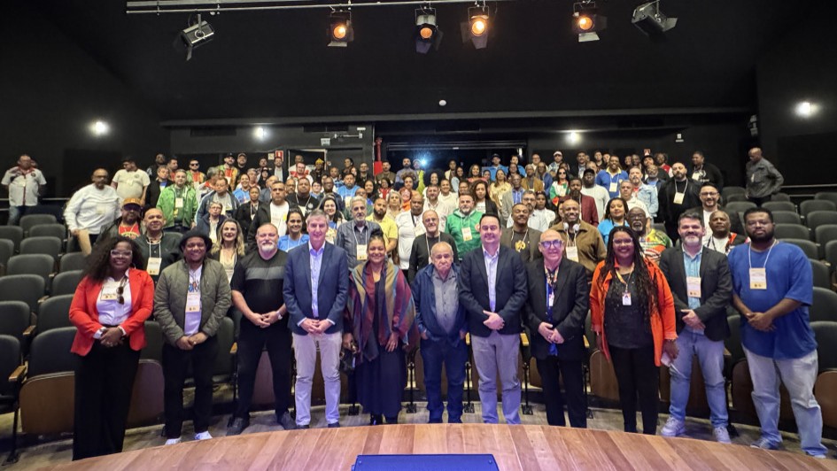 A imagem mostra uma foto de grupo tirada em um auditório com iluminação de palco. Há aproximadamente 100 pessoas presentes, distribuídas entre o palco (na frente) e as poltronas do auditório (ao fundo). Na primeira fileira, à frente, estão cerca de 20 pessoas em pé, possivelmente autoridades, organizadores ou convidados de destaque. Entre elas, há homens e mulheres de diferentes idades e etnias, vestidos de forma formal e semiformal, alguns com crachás de identificação pendurados no pescoço.