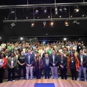 A imagem mostra uma foto de grupo tirada em um auditório com iluminação de palco. Há aproximadamente 100 pessoas presentes, distribuídas entre o palco (na frente) e as poltronas do auditório (ao fundo). Na primeira fileira, à frente, estão cerca de 20 pessoas em pé, possivelmente autoridades, organizadores ou convidados de destaque. Entre elas, há homens e mulheres de diferentes idades e etnias, vestidos de forma formal e semiformal, alguns com crachás de identificação pendurados no pescoço.