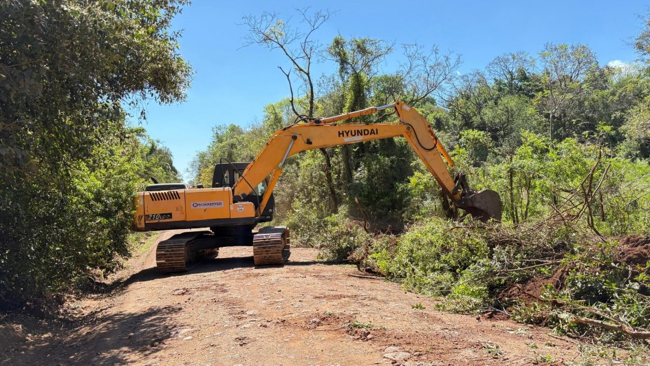 Foto de escavadeira trabalhando numa área aberta, de vegetação verde numa estrada de terra.