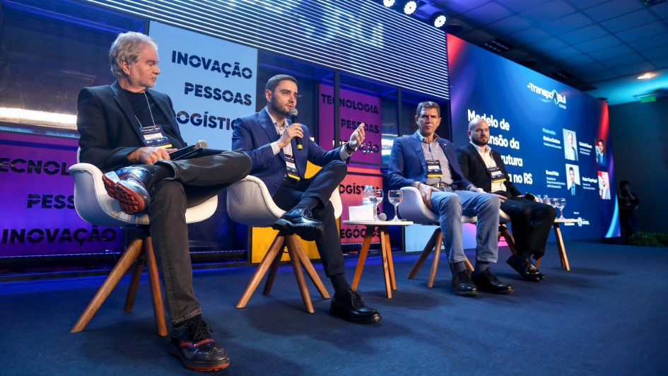 Na imagem, quatro homens estão sentados em cadeiras brancas sobre um palco, participando de um painel da TranspoSul. O vice-governador do Rio Grande do Sul, Gabriel Souza, aparece em destaque ao centro, vestindo terno azul e segurando um microfone enquanto fala ao público. Atrás, há painéis coloridos com palavras como "Inovação", "Pessoas" e "Logística". O ambiente é iluminado com luzes de palco, e uma tela grande exibe informações sobre o evento.
