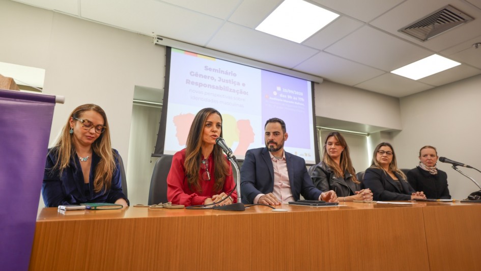 Cinco mulheres e um homem compõem uma mesa de debates. Uma das mulheres fala ao microfone, enquanto os demais escutam. Ao fundo, há um painel com a marca do Seminário Gênero, Justiça e Responsabilização. 