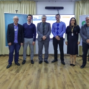 Seis pessoas estão em pé lado a lado, posando para foto em um ambiente interno com cortinas claras ao fundo e piso de madeira. À esquerda, há totens coloridos com a frase “Ganha Ganha Farroupilha”. Ao fundo, um telão exibe informações digitais. Entre os presentes, homens e mulheres vestem roupas formais e sociais, transmitindo caráter institucional e profissional ao evento.