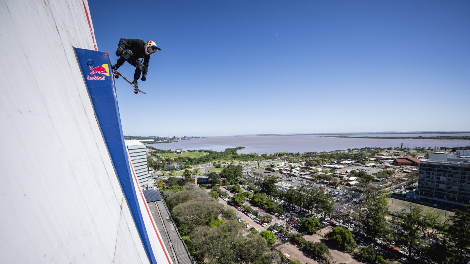 A imagem mostra um skatista realizando uma manobra aérea ousada na lateral de um prédio alto, que exibe a marca Red Bull. O atleta está suspenso no ar com o skate, evidenciando habilidade e coragem. Ao fundo, vê-se uma ampla paisagem urbana com diversos prédios, áreas arborizadas e um grande corpo d’água, tudo sob um céu azul claro.