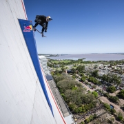 A imagem mostra um skatista realizando uma manobra aérea ousada na lateral de um prédio alto, que exibe a marca Red Bull. O atleta está suspenso no ar com o skate, evidenciando habilidade e coragem. Ao fundo, vê-se uma ampla paisagem urbana com diversos prédios, áreas arborizadas e um grande corpo d’água, tudo sob um céu azul claro.