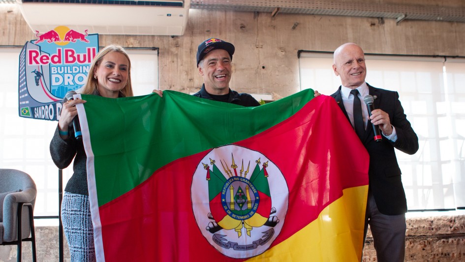 Esta é uma fotografia de três pessoas, dois homens e uma mulher, em pé em um ambiente interno, segurando uma grande bandeira do Rio Grande do Sul.

A bandeira, que domina o centro da imagem, é verde, vermelha e amarela, com o brasão do estado no meio. Os três sorriem e olham para a câmera.

À esquerda, uma mulher de cabelo loiro, usando um suéter escuro sobre uma camisa branca e uma saia xadrez cinza, segura a bandeira.

No centro, um homem com um boné preto e uma camiseta preta (o skatista Sandro Dias), também sorri e segura a bandeira.

À direita, um homem calvo, de terno preto e gravata, segura a bandeira com uma mão e um microfone com a outra, como se estivesse prestes a falar.

O fundo é uma parede de concreto aparente e um teto com vigas e um ar-condicionado visíveis. No canto superior esquerdo, há um banner vertical da Red Bull. A luz natural entra pelas janelas no fundo.