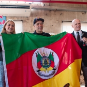 Esta é uma fotografia de três pessoas, dois homens e uma mulher, em pé em um ambiente interno, segurando uma grande bandeira do Rio Grande do Sul.

A bandeira, que domina o centro da imagem, é verde, vermelha e amarela, com o brasão do estado no meio. Os três sorriem e olham para a câmera.

À esquerda, uma mulher de cabelo loiro, usando um suéter escuro sobre uma camisa branca e uma saia xadrez cinza, segura a bandeira.

No centro, um homem com um boné preto e uma camiseta preta (o skatista Sandro Dias), também sorri e segura a bandeira.

À direita, um homem calvo, de terno preto e gravata, segura a bandeira com uma mão e um microfone com a outra, como se estivesse prestes a falar.

O fundo é uma parede de concreto aparente e um teto com vigas e um ar-condicionado visíveis. No canto superior esquerdo, há um banner vertical da Red Bull. A luz natural entra pelas janelas no fundo.
