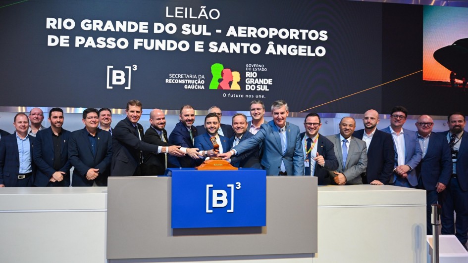 Grupo de autoridades em pé atrás de um púlpito, durante um evento oficial sobre aeroportos no Rio Grande do Sul. Ao fundo, um painel com informações do leilão e o logotipo da bolsa de valores B3.