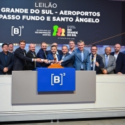 Grupo de autoridades em pé atrás de um púlpito, durante um evento oficial sobre aeroportos no Rio Grande do Sul. Ao fundo, um painel com informações do leilão e o logotipo da bolsa de valores B3.