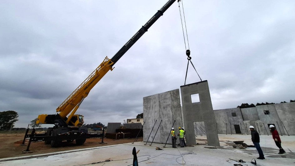 A imagem mostra o canteiro de obras de uma escola, com um guindaste amarelo erguendo uma grande parede de concreto pré-moldado. Ao fundo, há outras estruturas semelhantes já instaladas. Seis trabalhadores com capacetes e coletes de segurança acompanham a operação no solo. O ambiente é aberto, em um terreno amplo, com o céu nublado.