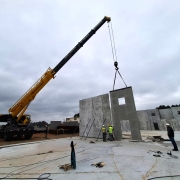 A imagem mostra o canteiro de obras de uma escola, com um guindaste amarelo erguendo uma grande parede de concreto pré-moldado. Ao fundo, há outras estruturas semelhantes já instaladas. Seis trabalhadores com capacetes e coletes de segurança acompanham a operação no solo. O ambiente é aberto, em um terreno amplo, com o céu nublado.