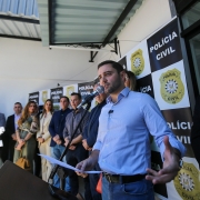 Fotografia de um evento oficial da Polícia Civil do Rio Grande do Sul. Em primeiro plano, um homem — identificado como o vice-governador Gabriel Souza — está falando ao microfone e segurando papéis, aparentemente fazendo um discurso. Ao fundo, há um painel institucional com o logotipo da Polícia Civil e a inscrição “POLÍCIA CIVIL”. Outras pessoas estão posicionadas ao lado e atrás do orador, indicando uma cerimônia formal, possivelmente a inauguração de uma delegacia. O ambiente é interno e bem iluminado, com foco na solenidade e na presença de autoridades.