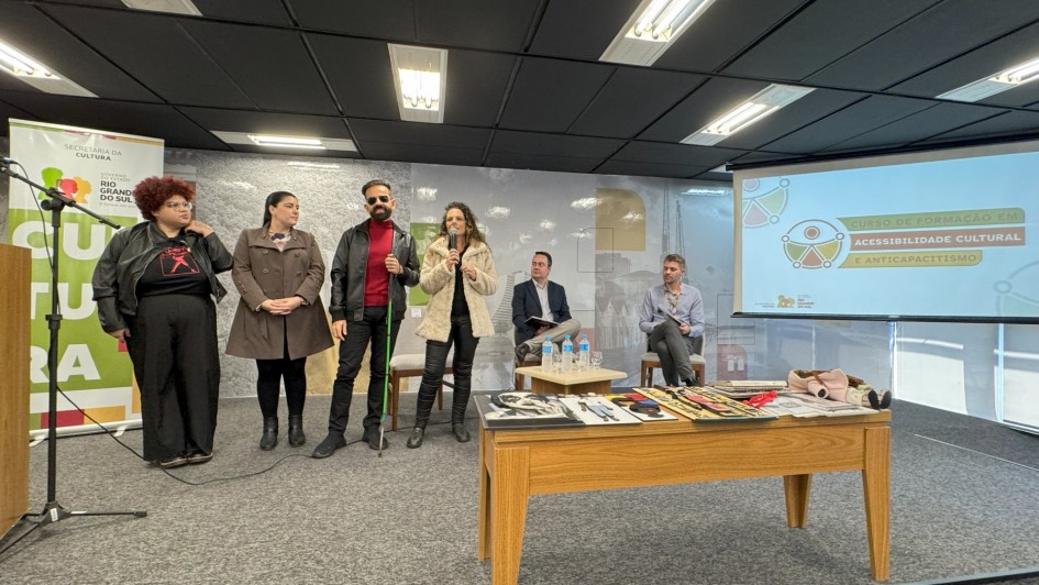 Na foto, cinco pessoas estão em pé no palco de um auditório. À esquerda, duas mulheres e um homem com bengala verde estão em destaque. A consultora Marina Baffini fala ao microfone. À direita, duas pessoas estão sentadas em cadeiras. Um banner da Secretaria da Cultura do RS aparece no canto esquerdo e um telão exibe os dizeres “Curso de Formação em Acessibilidade Cultural e Anticapacitismo”.