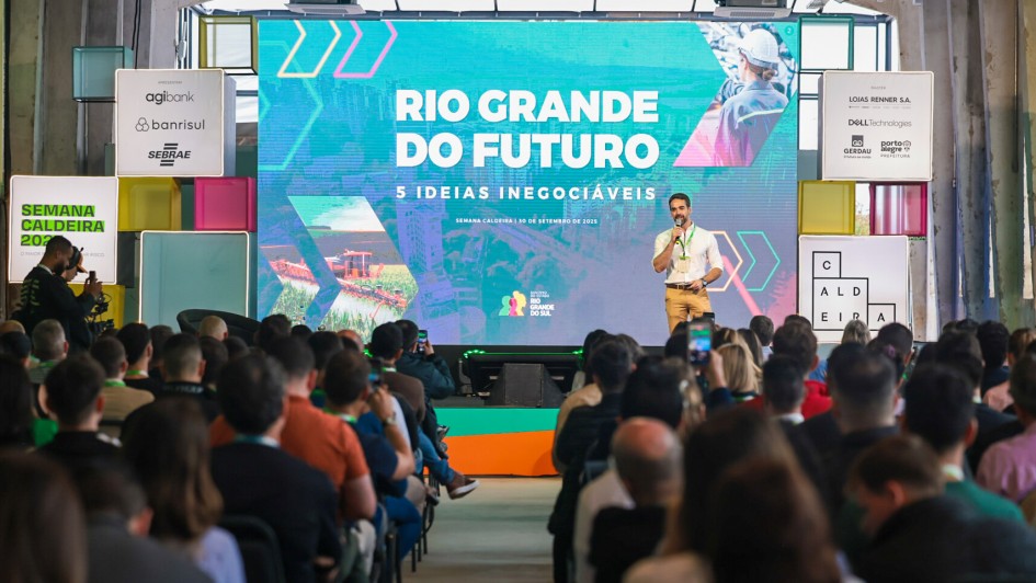 A imagem mostra o governador em um palco durante um evento realizado em um espaço amplo com estilo industrial. Ao fundo, há uma grande tela exibindo os dizeres “RIO GRANDE DO FUTURO” e “5 IDEIAS INEGOCIÁVEIS”. O público está sentado e atento. Banners e sinalizações ao redor do palco incluem o nome “SEMANA CALDEIRA”.