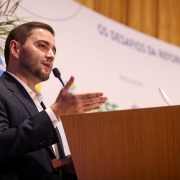 Um homem de pele clara e barba curta aparece em primeiro plano, falando ao microfone em um púlpito de madeira. Ele veste terno escuro e camisa branca, gesticulando com a mão direita aberta. Ao fundo, há uma bandeira do Brasil parcialmente visível e um painel branco com logotipos e a frase “OS DESAFIOS DA REFORMA TRIBUTÁRIA” parcialmente legível. À esquerda, ramos de uma planta verde completam a cena.