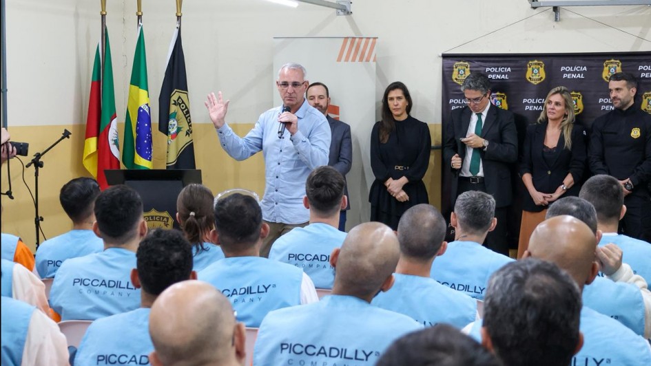 Na imagem, o secretário Jorge Pozzobom fala ao microfone para um grupo de pessoas usando coletes azuis com a inscrição Piccadilly. Atrás dele, ao lado de autoridades e convidados, estão as bandeiras do Rio Grande do Sul, do Brasil e da Polícia Penal. 