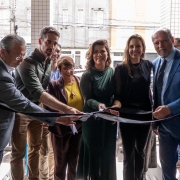 Um grupo de sete pessoas está reunido em uma cerimônia de inauguração, cortando uma fita preta com laço branco. Todos estão sorrindo e posicionados próximos uns dos outros. A maioria está vestindo trajes formais — ternos e vestidos. O local parece ser a entrada de um prédio moderno, com revestimento de pastilhas claras. Ao fundo, pela porta de vidro, é possível ver uma rua movimentada, com prédios antigos, lojas e fiação elétrica. A cerimônia parece contar com a presença de autoridades ou convidados importantes.