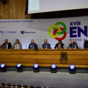 A imagem mostra um auditório com um painel de debates composto por sete pessoas sentadas atrás de uma longa mesa de madeira, sobre um palco. Ao fundo, há um grande painel com o logotipo do evento “XVIII ENAT – Porto Alegre” e o tema “Os desafios da reforma tributária”.