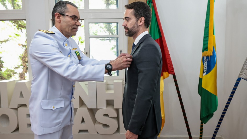 Em ambiente interno iluminado por luz natural, um militar em uniforme branco da Marinha do Brasil prende uma medalha na lapela do paletó de um homem trajando terno escuro. Ao fundo, vê-se parcialmente um painel com letras brancas e duas bandeiras: a do Brasil (verde, amarela e azul) e a do Rio Grande do Sul (vermelha, verde e amarela). A cena retrata o momento da entrega da Medalha Mérito Tamandaré ao governador Eduardo Leite.