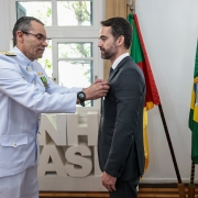 Em ambiente interno iluminado por luz natural, um militar em uniforme branco da Marinha do Brasil prende uma medalha na lapela do paletó de um homem trajando terno escuro. Ao fundo, vê-se parcialmente um painel com letras brancas e duas bandeiras: a do Brasil (verde, amarela e azul) e a do Rio Grande do Sul (vermelha, verde e amarela). A cena retrata o momento da entrega da Medalha Mérito Tamandaré ao governador Eduardo Leite.