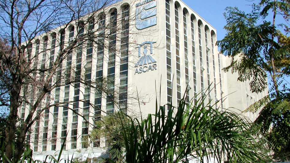 A imagem mostra um prédio de vários andares, com fachada clara e janelas altas e estreitas distribuídas em colunas verticais. A arquitetura possui um estilo moderno, com arcos nas extremidades superiores das janelas. Na lateral do edifício, estão visíveis dois logotipos grandes: um com a palavra "SULBRASILEIRA" e o outro com "ASCAR", ambos acompanhados de símbolos gráficos. O prédio está parcialmente coberto por árvores e vegetação na frente, e o céu ao fundo está claro e azul, indicando um dia ensolarado.