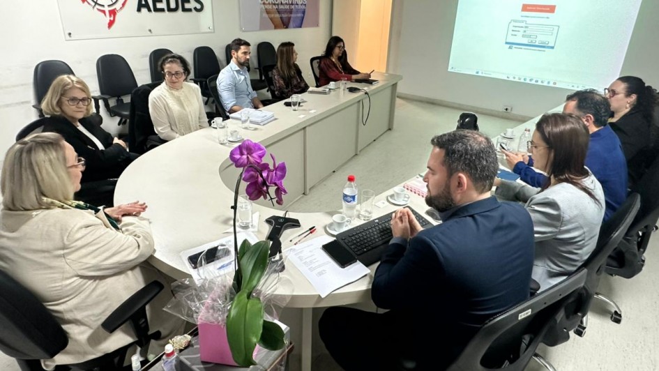 Imagem mostra sala de reunião da SES com uma mesa em formato de U com secretária numa das pontas e outras pessoas dispostas em outros pontos da mesa. 