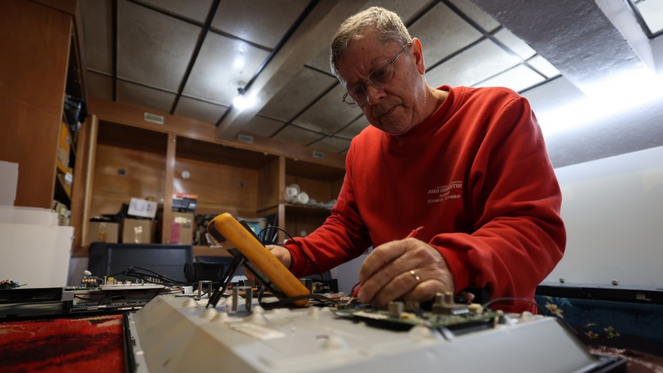 A foto mostra um homem concentrado no conserto de um equipamento eletrônico. Ele usa um multímetro para fazer medições em uma placa de circuito sobre uma bancada de trabalho. Veste moletom vermelho e óculos.