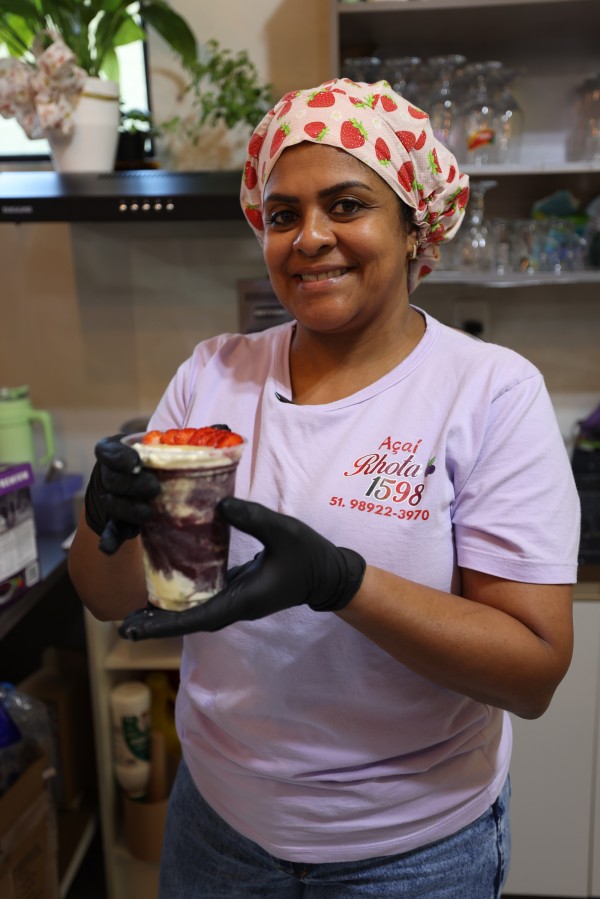 Uma mulher sorri segurando um copo de açaí bem decorado com pedaços de morango no topo. Ela usa luvas pretas e uniforme lilás com o nome de sua empresa estampado, além de um lenço colorido na cabeça com estampas de morangos.