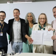 A imagem mostra um grupo de cinco pessoas reunidas em um ambiente interno, posando para uma foto durante um momento formal. No centro, duas pessoas seguram um documento aberto.
