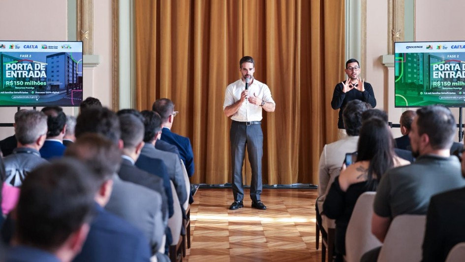 Cena de um auditório, onde Eduardo Leite aparece em pé, no centro, falando ao microfone diante de uma plateia. Ele veste camisa branca e calça cinza. À sua direita, um intérprete de Libras traduz seu discurso. Dois telões exibem o título do programa “Porta de Entrada – Fase 2”, com destaque para o valor de R$ 150 milhões.