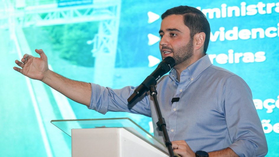 Homem de camisa azul claro fala ao microfone em um palco, gesticulando com a mão. Atrás dele, há uma tela grande com texto em português e a imagem de uma estrada. Parece ser uma apresentação ou conferência.